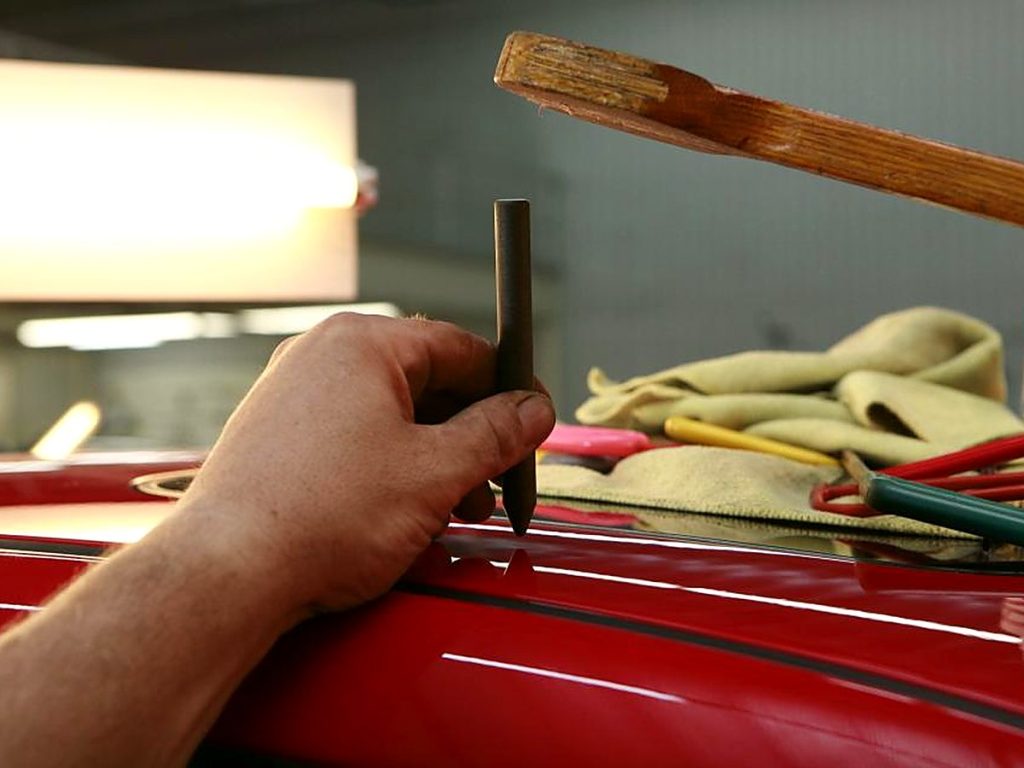 Een hand werkt met hamer en pen een deukje uit rode lak